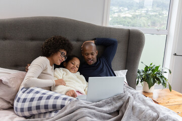 Diverse family with daughter watching laptop on bed under window, daughter wearing white robe © wavebreak3