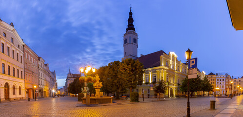 Fototapeta premium Evening view of the market square in Swidnica, Poland with town hall tower, fountain, and historic buildings illuminated under blue hour sky.
