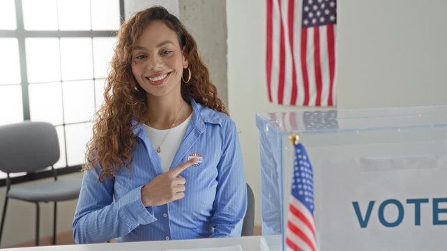 Woman points finger to voted sticker on chest beside clear ballot box and american flag at voting table inside building; pride civic duty.