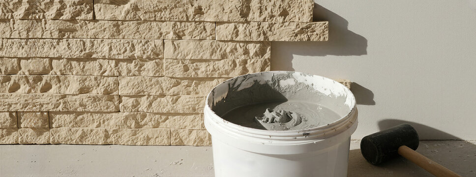Bucket with mortar and rubber mallet near stone wall. Construction site with tiling material for wall cladding. Banner with copy space