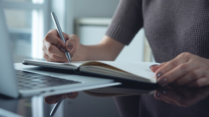 Woman writing in notebook with laptop on table during working at home office, doing online...
