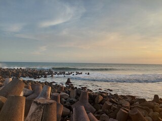 Obraz premium Golden hour seascape featuring gentle ocean waves, a rocky shoreline, and concrete breakwater structures under a dramatic cloudy sky.