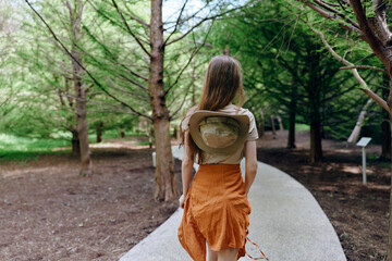 Fototapeta premium Woman walking on a park path with hat on her back, orange skirt and casual top, surrounded by trees and greenery. Outdoor stroll, summer fashion and peaceful nature escape.