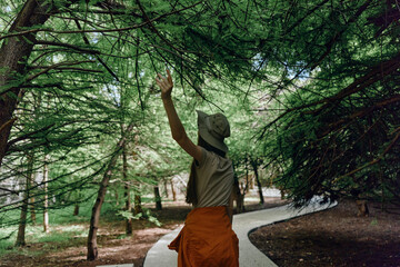 Fototapeta premium Woman hiking on a winding forest path, reaching up to tree branches on a nature trail. Tall trees and dappled light surround her, wearing a hat and jacket tied around the waist.