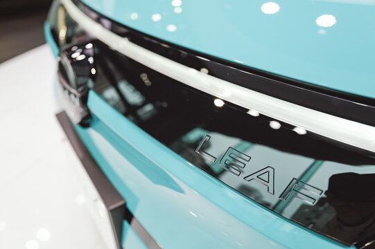 Close-up of the Leaf emblem on a Nissan electric vehicle in a showroom. the "LEAF" model name embossed on the sleek black trim of a vibrant light blue Nissan electric car.