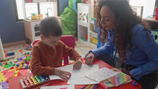 Boy drawing with pencil and hand on paper at a small red table inside a building playroom; learning concentration.