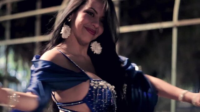 Beautiful Latina woman dancing belly dance, moving her hands and twirling, wearing a sexy blue dress, dancing for the camera, wearing makeup. Dancing and smiling, with sensuality and joy.