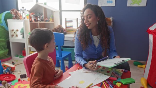 Boy student draws with crayon while teacher holds a clipboard and points pen in preschool building; calm concentration.