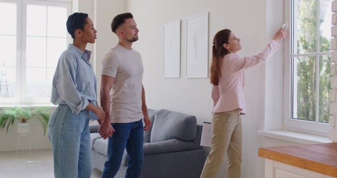 Realtor woman showcasing modern property to young multiethnic spouses, explain benefits, talking about amenities, standing together in unfurnished, spacious living room. Bank loan, tenancy