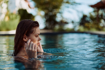 Fototapeta premium Young woman relaxes in a sunlit pool, fingers resting on her cheeks as gentle water ripples frame a serene moment