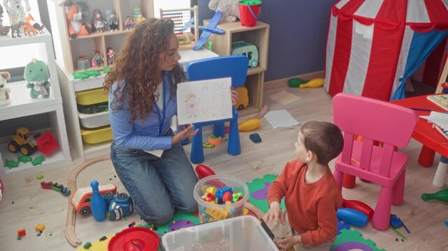 Woman teacher kneeling in preschool playroom holds up abc drawing while little boy points finger to paper among scattered toys in building; curiosity focused learning.