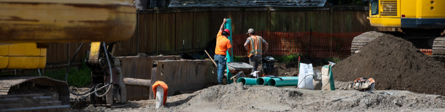Stormwater system pipe installation in progress, construction workers with 6 in green PVC pipes standing above excavated dirt trench on infrastructure work site
