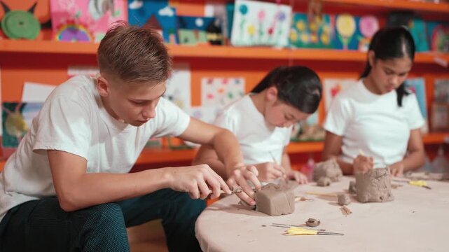 high school teenager using carving tool working at clay at pottery workshop. Group of happy diverse children working or modeling cup of clay with dough and equipment scatter around. Edification