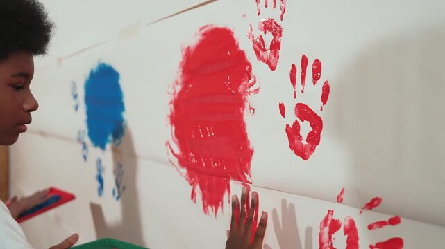 Diverse happy high school girl and african boy paint the wall with water color in red and blue or contrast color by using their hands in art lesson. Side view. Creative activity concept. Edification
