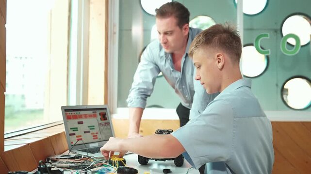 Caucasian teacher pointing at laptop screen and giving advise for programming system or coding prompt while smart high school boy listen and use equipment to repair robot at STEM classroom