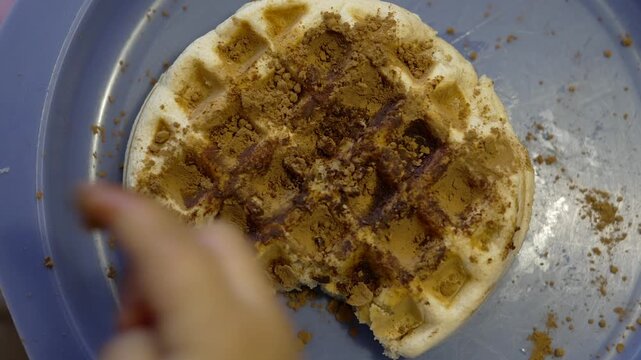Top view of a round waffle on a blue plate covered with chocolate powder while a child&rsquo;s hand picks and tastes only the topping without eating the waffle. Concept of picky eating,