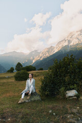 Fototapeta premium A woman sits on a rock in an open meadow with distant mountains, calm and reflective. A lifestyle portrait showing outdoor serenity and nature inspired mood.
