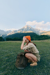 Fototapeta premium Young woman crouching in a grassy field with a backpack and orange hat, wearing casual wear. A mountain range rises in the background, conveying outdoor lifestyle and travel adventure.