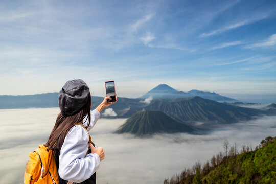 Young woman tourist with backpack taking a photo with phone of the Mount Bromo crater and sea of mist
