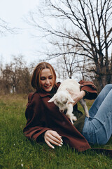Fototapeta premium Young woman with braided hair lies on grass holding a white baby goat in her arms during a relaxed outdoor lifestyle moment in early spring.