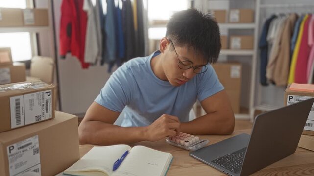 Man writing in notebook beside laptop and stacked shipping boxes, hand holding pen and touching pillbox organizer in studio; focused entrepreneurship.