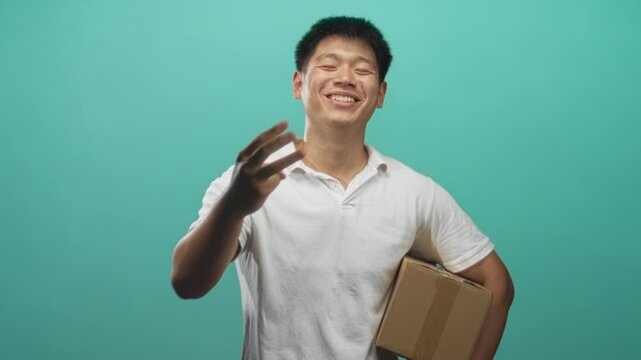Young chinese man holding cardboard box with a fist pump and pointing gesture in studio; joy success delivery.