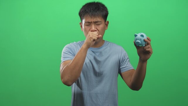 Young man holding a blue piggy bank and coughing into his fist while looking distressed against a green studio screen; financial anxiety.