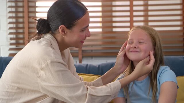 Mother talking and touch of face her daughter while sitting together on sofa, showing caring parenting and communication at home