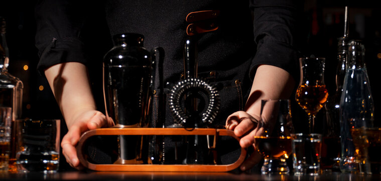 Bar tools kit preparation with shaker and strainer by woman bartender on dark counter. Professional mixology equipment and cocktail preparation workspace concept in modern restaurant setting.