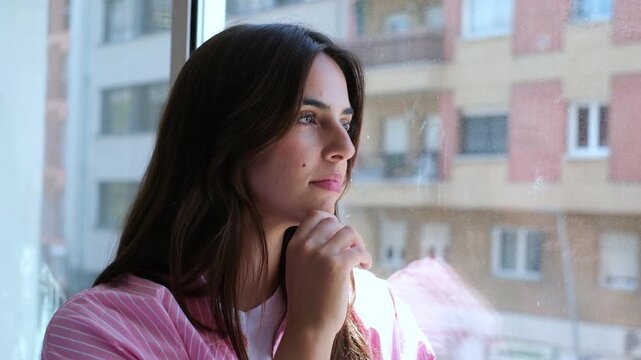 A thoughtful young woman gazing out her apartment window at the city. She appears pensive, lost in her thoughts, reflecting on life and possibilities.