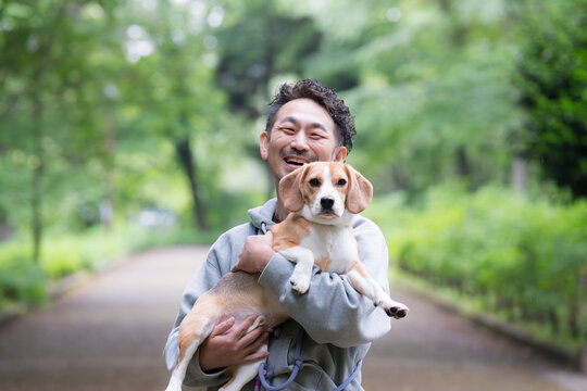 新緑背景で愛犬のビーグルを抱く男性の笑顔、公園で過ごす幸せなお散歩時間のカメラ目線クローズアップ