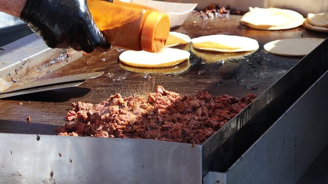Taco meat cooking on an open grill and being seasoned while flour shells are warming in the back.