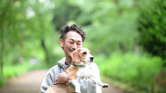 新緑背景で愛犬のビーグルを抱く男性の笑顔、公園で過ごす幸せなお散歩時間のカメラ目線クローズアップ動画	スローモーション