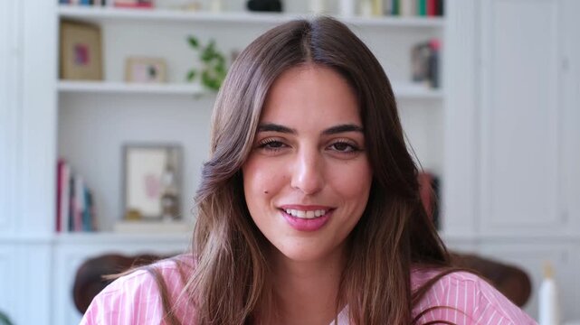 A beautiful young woman with long brown hair smiles genuinely at the camera. Her radiant expression conveys happiness and positive emotion in a bright, comfortable home setting.