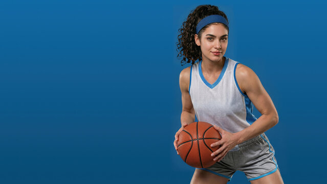 Young woman in silver basketball attire grips a ball in a ready stance against a blue studio background, with a headband. Copy space.