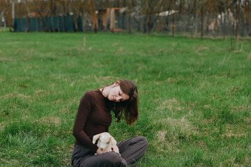 Fototapeta premium A woman in a brown sweater sits on the grass, cradling a gentle goat kid in her arms in a wide field. A calm lifestyle moment illustrating connection with animals, nature, and outdoor living.