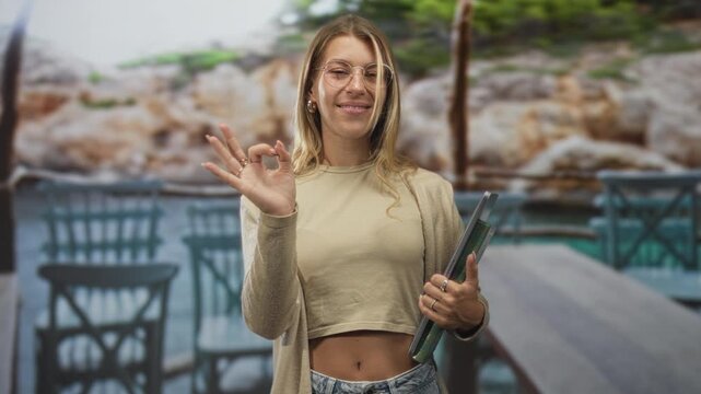 Woman holding green binder and making ok sign with right hand at seaside cafe terrace; satisfaction confidence.