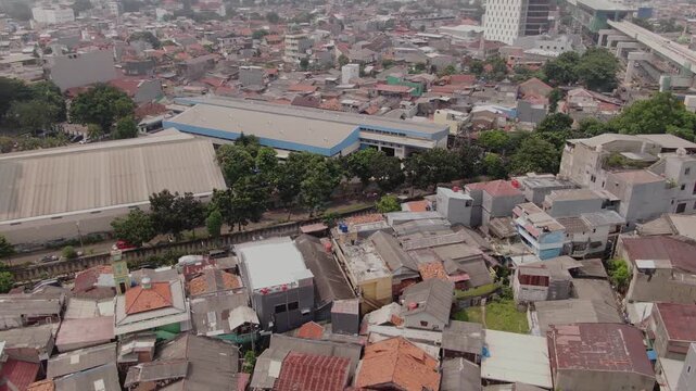 Aerial view of industrial warehouse facility next to dense residential housing area. High angle shot of urban landscape featuring logistics building, trees, and crowded neighborhood in megacity