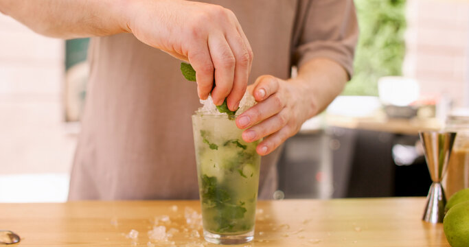 Garnishing mojito. Serving cocktail. Unrecognizable barman hand adding fresh mint sprig on natural ice cool fruit drink at professional bar counter.