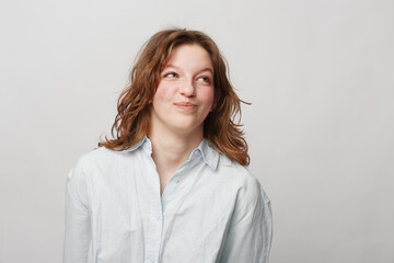 Obraz premium Young woman with curly hair wearing white shirt looks up with playful smile on light background. Concept of casual style, positive emotions, and natural beauty in studio portrait.