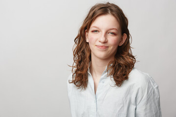 Obraz premium Young woman with curly brown hair smiles gently wearing a white shirt against a plain light background. Portrait of natural beauty, casual style, and confident expression.