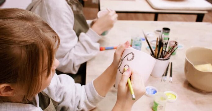 Craft education. Diy art. Two talented girls enjoying handmade pottery hobby artist painting on ceramic cup using pencil indoor.
