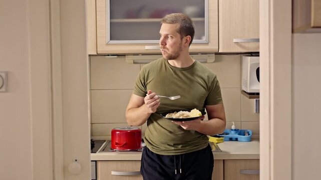 A young man stands relaxed in his modern home kitchen, eating a simple meal from a black plate with a fork. This scene depicts an everyday domestic routine and the comfort of home life.