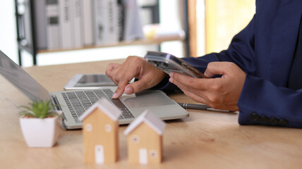 Business professional using smartphone and laptop at desk with house models symbolizing real estate investment, property management, and financial planning in a modern digital workspace environment. © Siwaporn