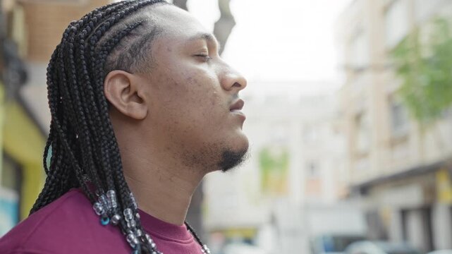 Man in profile with braided hair and beads, face visible and eyes closed on street; quiet reflection serenity.
