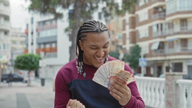 Man with braided hair holding a fan of banknotes, smiling and pumping his fist in a city street; joy wealth celebration.
