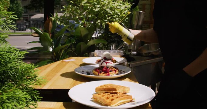 Waffles serving. Garnishing dessert. Unrecognizable chef man hand in gloves decorating sauce with whipped cream berries on white plate squeeze bottle in cafe outdoor.