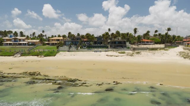Luxury beachfront villa with pool and palm garden facing sandy shoreline at Taiba beach in Sao Goncalo do Amarante municipality, Ceara state, Brazil, South America under bright daylight drone pullback