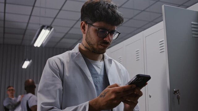 Concentrated Young Doctor With Tattoos on Arm Uses Smartphone to Study Critical Medical Reports and Lab Findings in Hospital Dressing Room After Work. Concept of Post Shift Clinical Analysis.