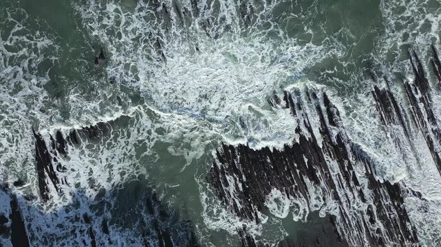 Aerial topdown view of waves breaking over dramatic flysch rock formations at Sakoneta beach, Basque Country, creating abstract coastal patterns and foaming sea texture